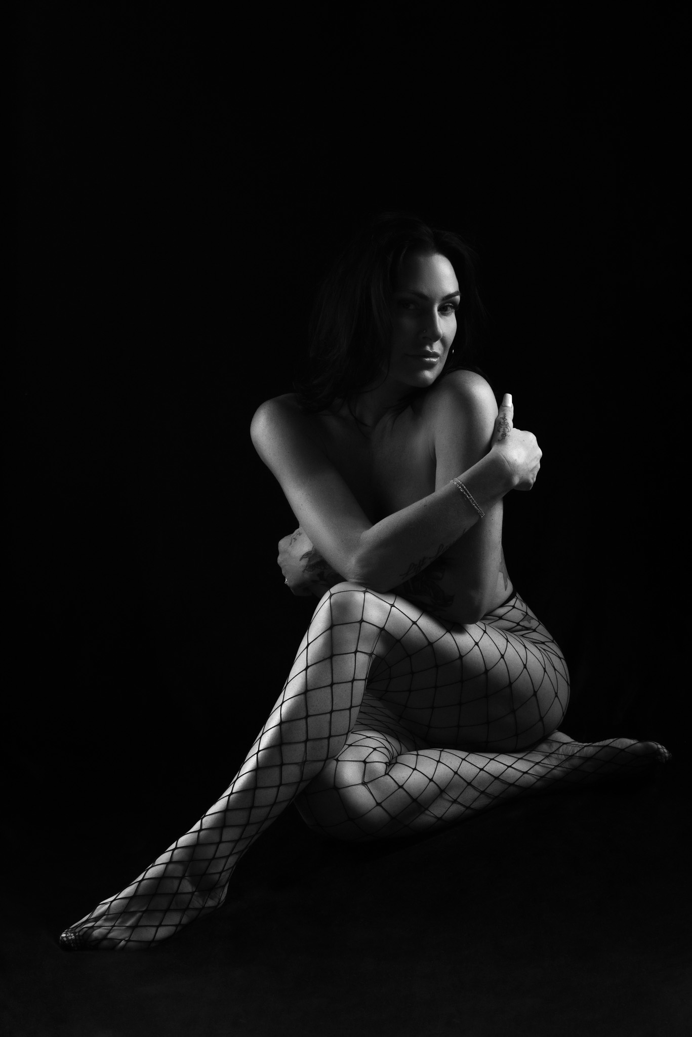 Dark and moody boudoir of a woman sitting cross legged giving herself a hug.